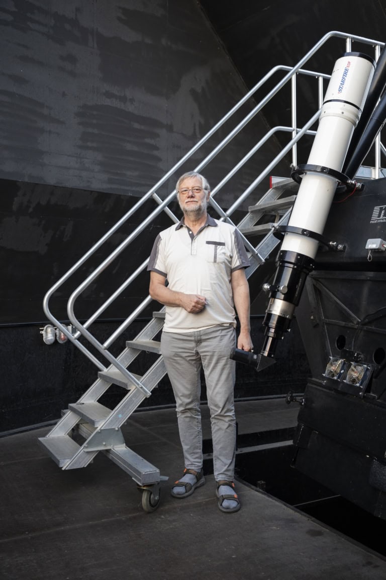 Der Rentner und Hobby-Astrologe Bernd Schröter steht in der Sternwarte. Im Hintergrund befindet sich die grau-schwarze Innenseite des Daches. Auf seiner rechten Seite ist ein weißes Teleskop, mit blau-roter Aufschrift ‚Starfire EDF‘, an einer schwarzen Halterung angebracht. Hinter ihm befindet sich eine silberne Metalltreppe, die entlang der schwarzen Halterung nach oben führt. Schröter trägt ein grau-weißes Hemd mit kurzen Ärmeln, eine graue lange Hose und hellblaue Socken mit grau-orangenen Sandalen. Er hat weiße Haare sowie einen weißen kurzen Vollbart und trägt eine Brille.