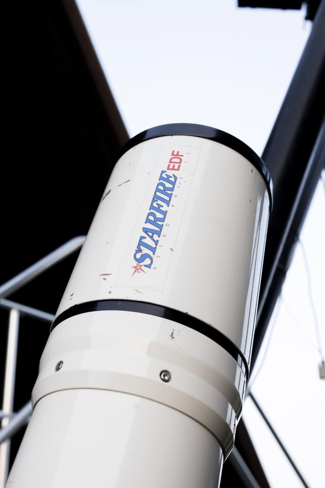 Der obere Teil eines weißen Teleskops ragt in Richtung grau-hellblauer Tageshimmel. Das Teleskop trägt die blau-rote Aufschrift ‚Starfire EDF‘. Im Hintergrund befindet sich verschwommen ein Ausschnitt der schwarzen Innenseite des Daches und ein Teil eines silbernen Gerüsts.