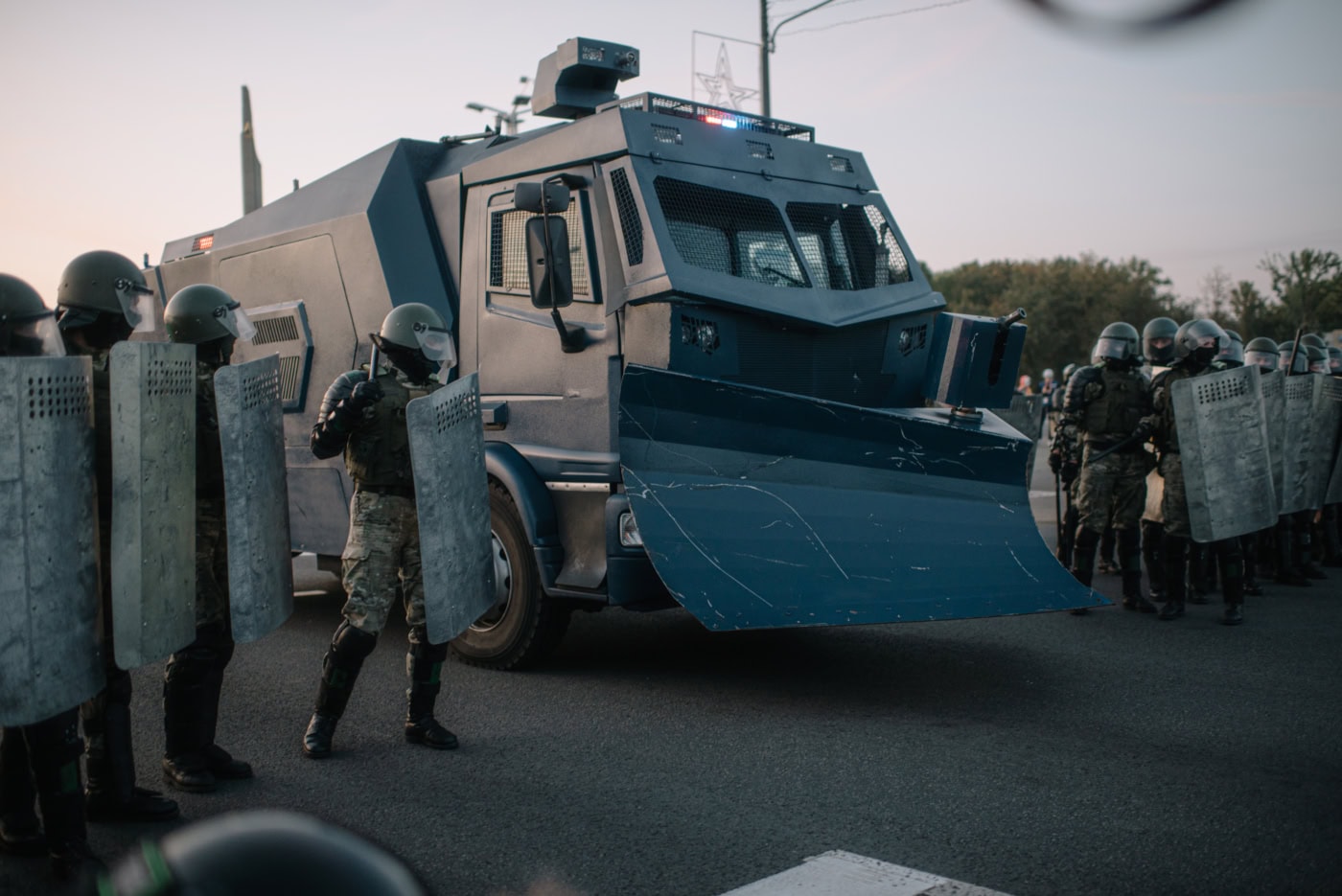 In der Dämmerung steht auf einer Straße ein bedrohlich wirkendes, gepanzertes Polizeifahrzeug. Links und rechts von dem Fahrzeug stehen uniformierte und gepanzerte Polizisten. Sie tragen helm, Schlagstock und ein Schutzschild.