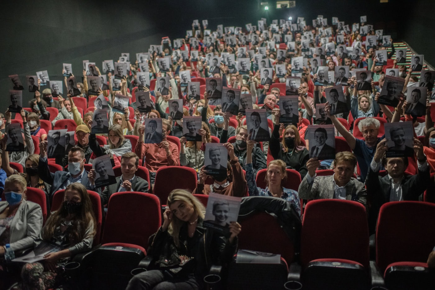 Menschen die in einem voll besetzten Kinosaal auf roten Sitzen sitzen, halten schwarz weiße Fotografien in die Luft, auf denen unterschiedliche Personen zu erkennen sind.