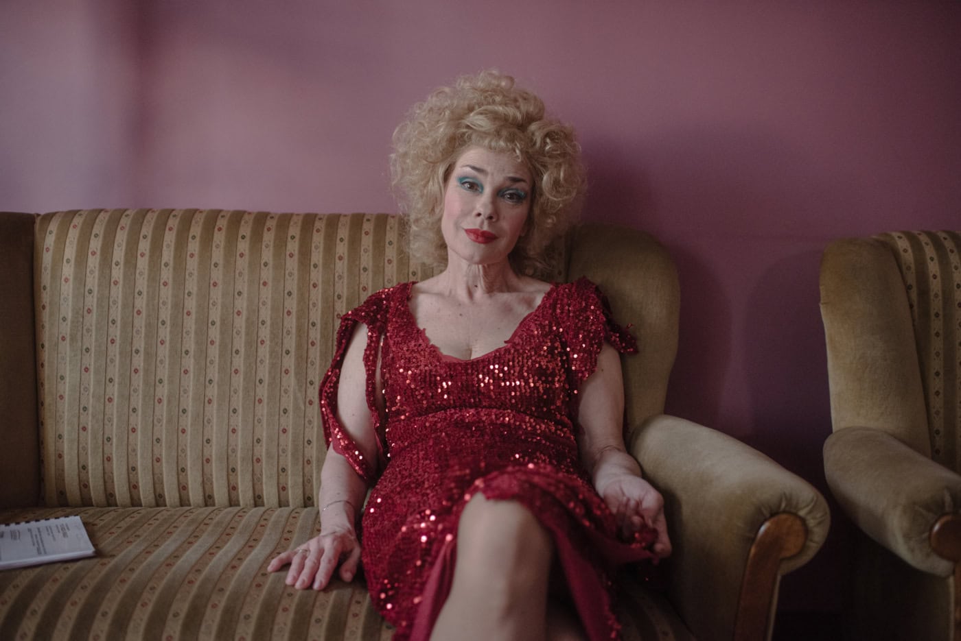 Ein ältere Frau sitzt auf einer Couch. Sie trägt starkes Make-Up, ein rotes glitzerndes Kleid und ihre blonden, lockigen Haare sind toupiert, fast als wären sie eine Perücke. Die Frau lächelt sanft in die Kamera. Die Wand hinter ihr ist rosa.