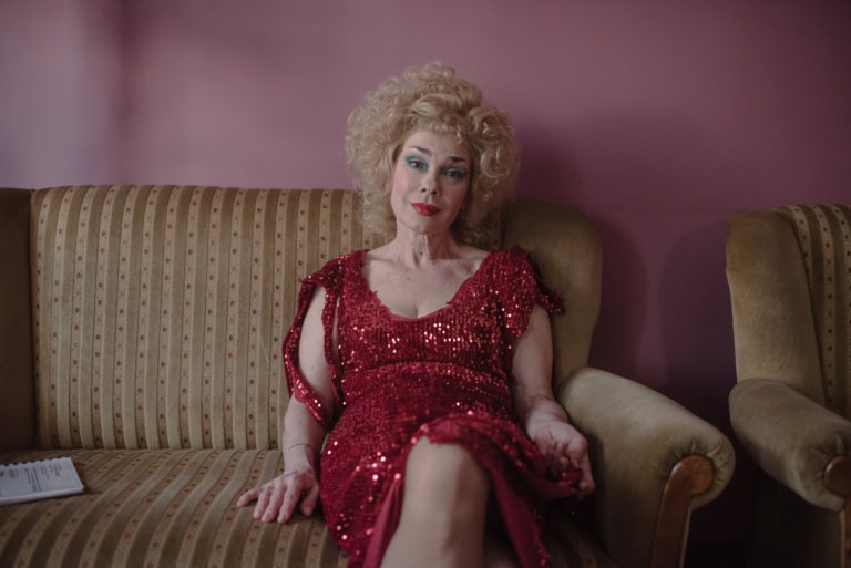 Ein ältere Frau sitzt auf einer Couch. Sie trägt starkes Make-Up, ein rotes glitzerndes Kleid und ihre blonden, lockigen Haare sind toupiert, fast als wären sie eine Perücke. Die Frau lächelt sanft in die Kamera. Die Wand hinter ihr ist rosa.