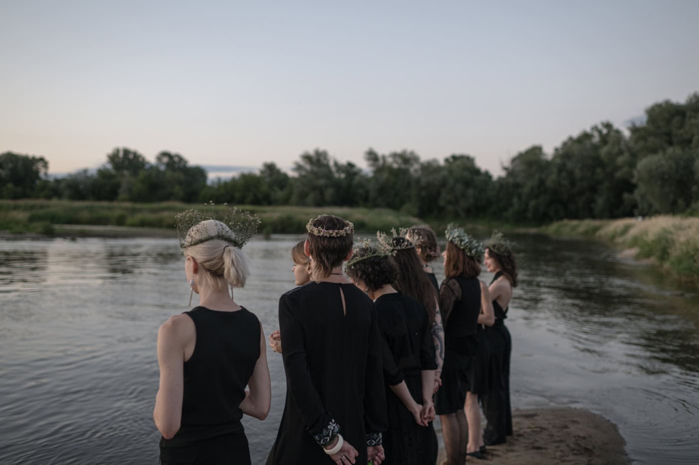 Acht junge Frauen stehen in der Dämmerung an einem Flussufer. Sie tragen schwarze Kleidung sowie Blumenkränze auf dem Kopf.