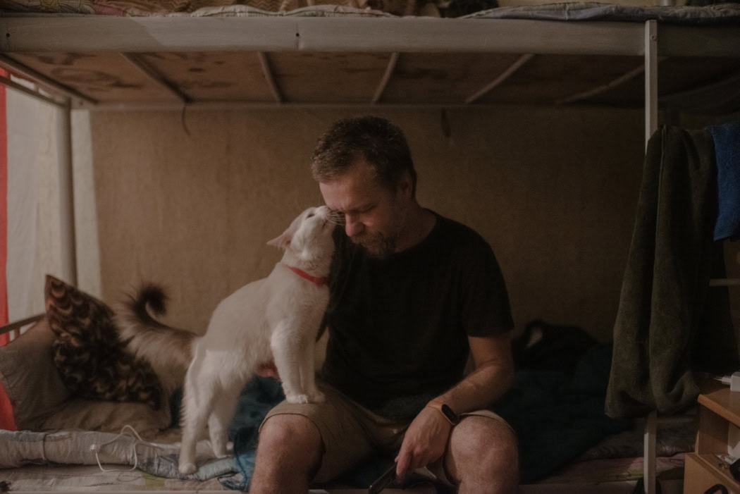 In der Bildmitte sitzt ein Mann auf dem unteren Teil eines Doppelstockbettes. Sanft guckt er nach links unten, während eine Katze sein Gesicht ableckt und mit den Vorderpfoten auf seinem Bein steht.