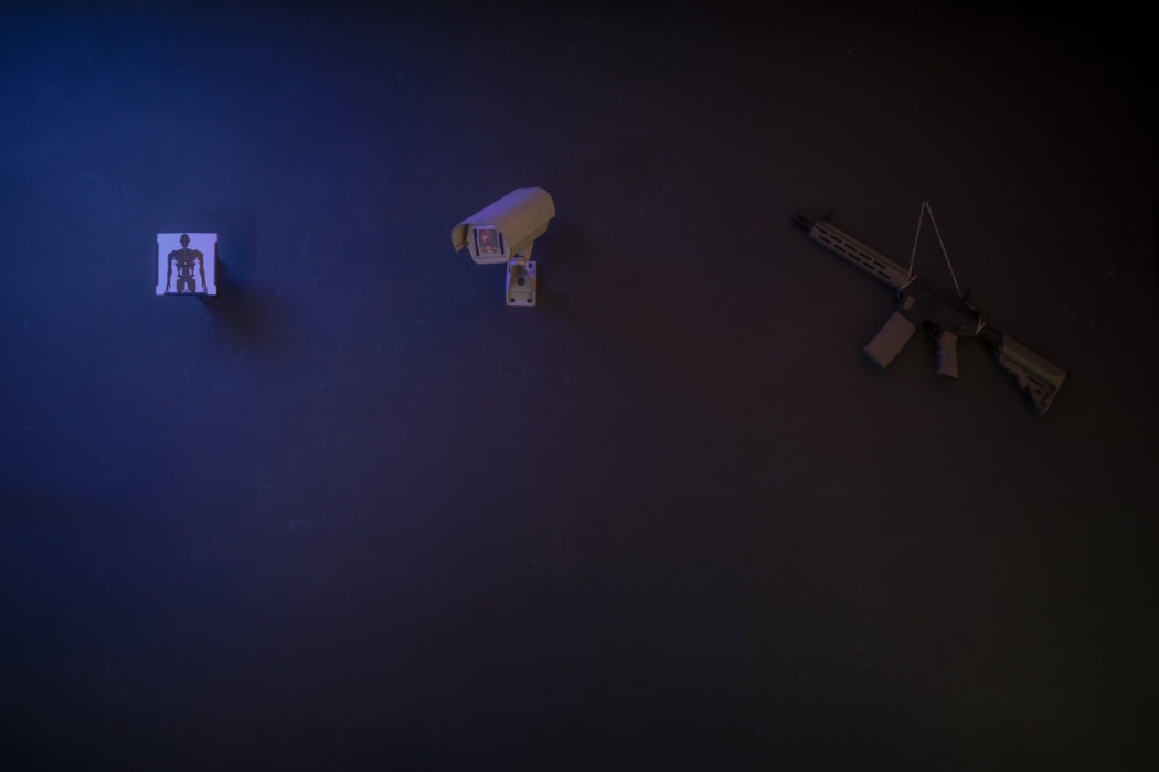 Zu sehen ist eine Wand in einem dunklen Raum. Es wirkt, als würde blauer Licht auf die Wand scheinen. An der Wand hängt von links nach rechts ein Stück Papier mit einer Silhouette drauf, die für Schussübungen verwendet wird, eine Überwachungskamera auf die eine Spielkarte geklebt wurde, sowie ein Gewehr. Es ist nicht zu erkennen ob dies echt ist oder ein Spielzeug.