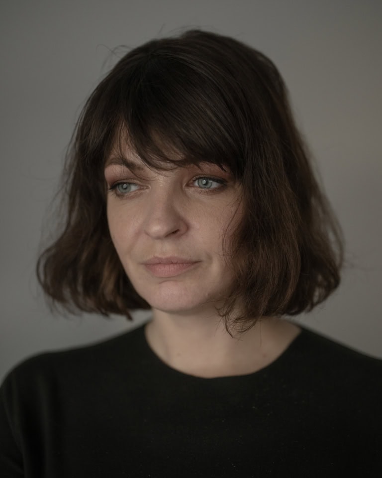 Ein Portrait einer Frau, etwa 30 Jahre alt. Ihre dunklen Haare gehen bis kurz über die Schultern. Sie schaut melancholisch links an der Kamera vorbei.