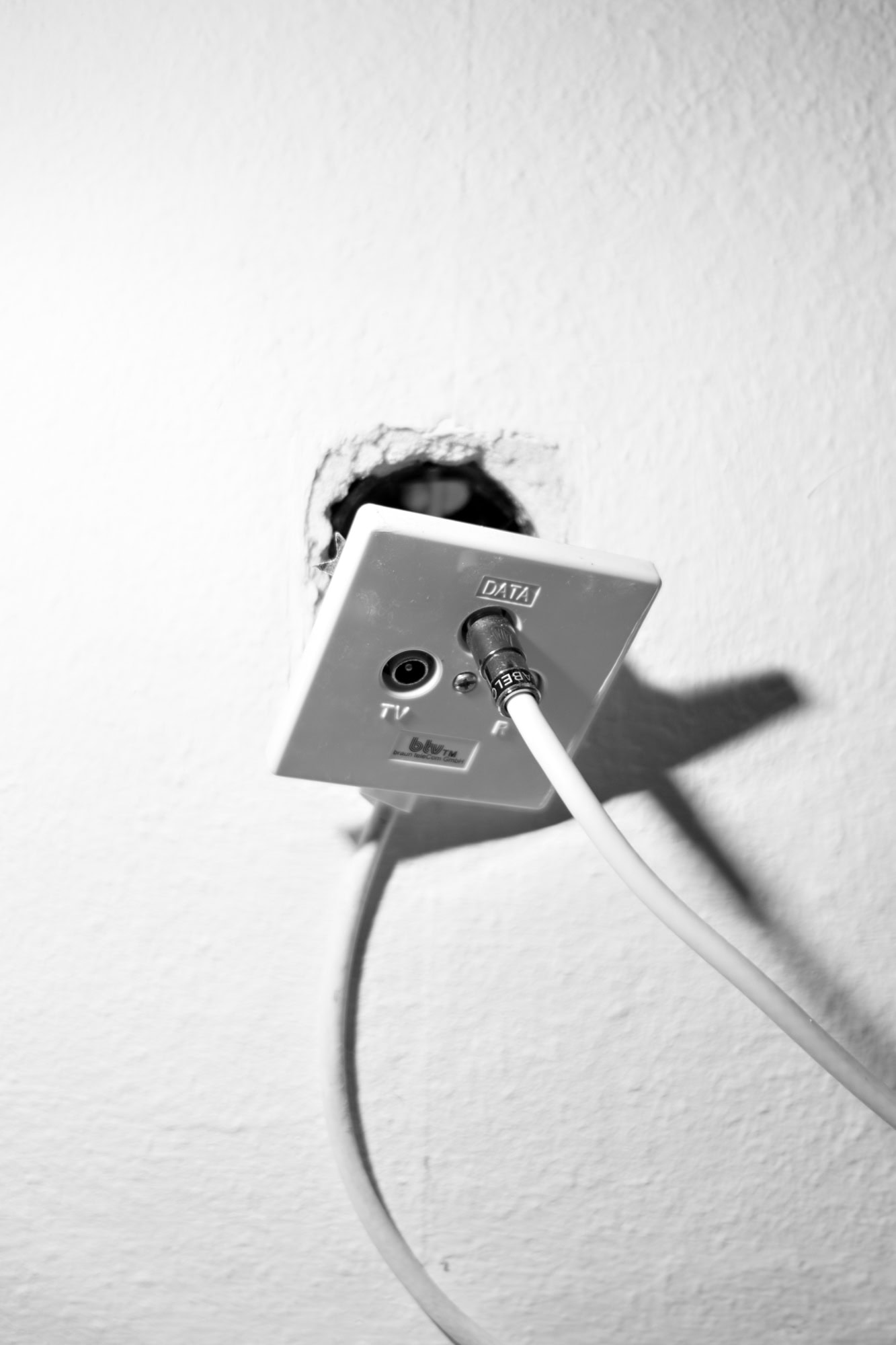 Eine weiße Wand mit einer Internet-Steckdose, an die zwei Kabel angeschlossen sind. Die Halterung ist aus der Wand rausgebrochen.