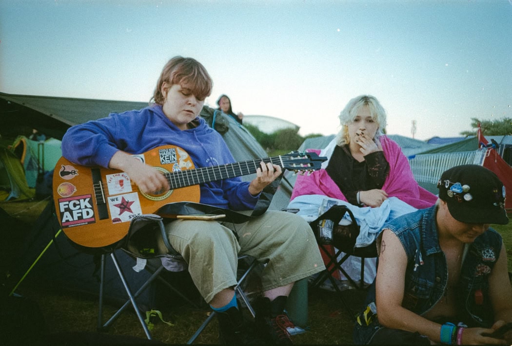 Drei Personen: Im Vordergrund spielt eine Person Gitarre, rechts daneben schaut eine Person auf ihr Handy, im Hintergrund steht eine weitere Person und raucht.