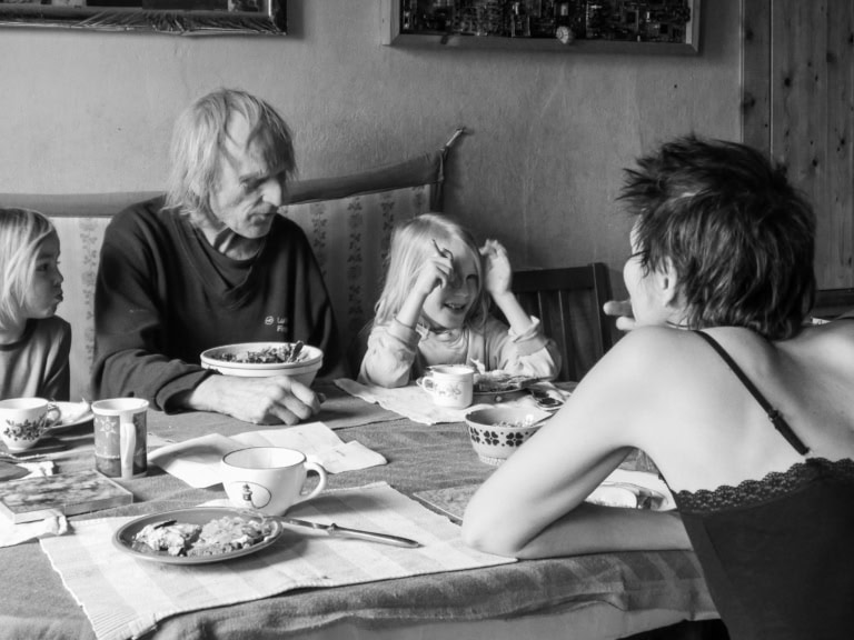 Schwarz/weiß Fotografie. Vier Menschen sitzen an einem Küchentisch und essen Frühstück. Der Tisch ist mit einer Tischdecke gedeckt. Zwei Kinder, ein alter Mann und eine junge Frau mit kurzen Haaren