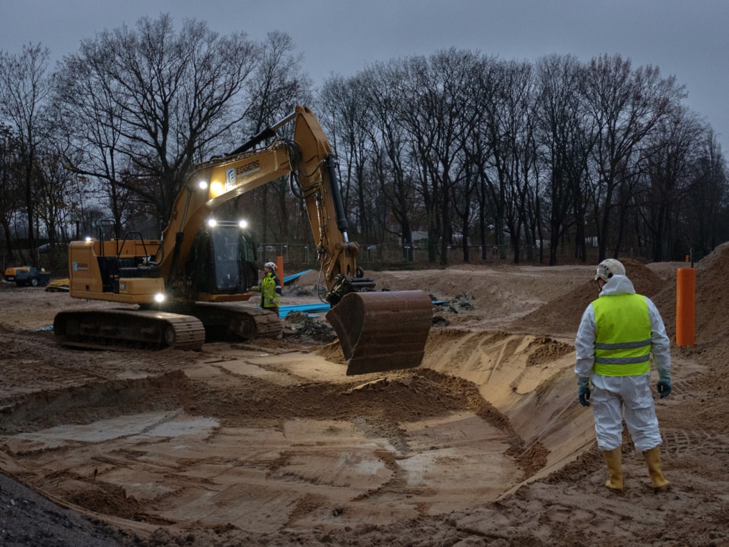 Ein Bagger hebt vorsichtig Erde aus, während zwei Arbeiter in Schutzanzügen und Warnwesten bei der Vorbereitung einer Bombenentschärfung auf einer Baustelle zuschauen. Die Szene spielt sich in einem bewaldeten Gebiet bei Tagesanbruch ab.