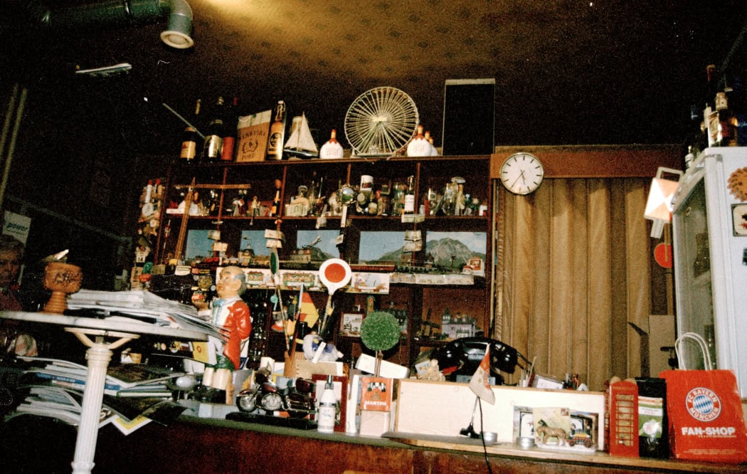 Theke mit zahlreichen kleinen Dekorationsobjekten, Flaschen und Souvenirs, darunter eine Figur eines Jungen in rotem Mantel, eine kleine grüne Kugelpflanze und ein roter Bayern München Fan-Shop Beutel.