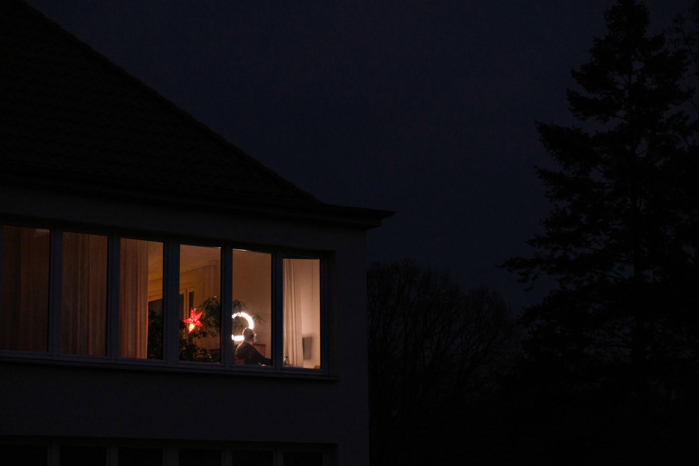 Ein farbiges querformatiges Bild. Es ist Nachts und dunkel, man sieht durch ein Fenster eine Person an einem Schreibtisch sitzen. Vor der Person steht ein Computer. Das Licht scheint im Wohnzimmer.