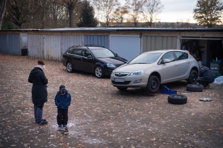 Eine Mutter und ihr kleiner Sohn warten, während deren Nachbar (nicht Garagennachbar) ihrem Auto Winterreifen aufzieht. Die Mutter dreht sich gerade eine Zigarette. Ort: Salzburger Str., 01279 Dresden ; Foto: Adam Beyer