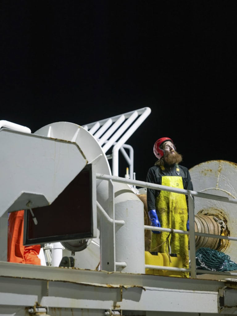 Es ist Nachts. Zu sehen ist ein Mann Bart, rotem Kopfschuss und gelber Latzhose. Sein blick geht aus dem Bild heraus und die Betrachtenden erkennen nicht auf was er schaut. Er steht auf einem Trawler. Die Szenerie ist ist gut ausgeleuchtet und die die Farben sehr kontrastreich.