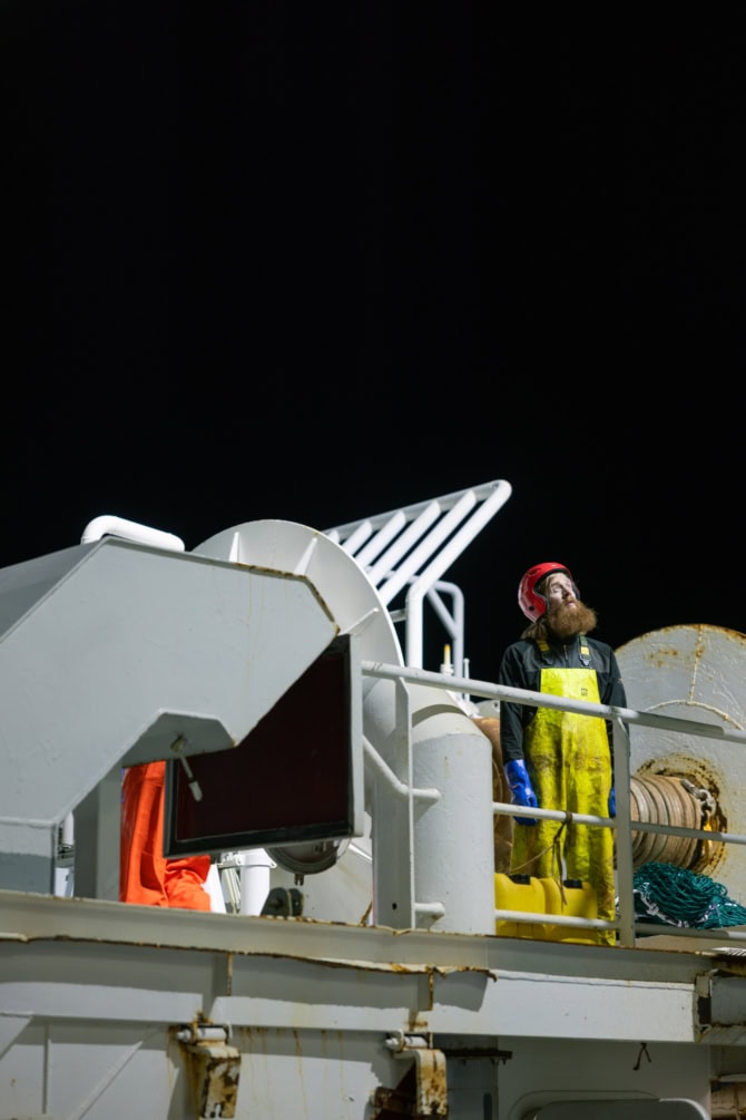 Es ist Nachts. Zu sehen ist ein Mann Bart, rotem Kopfschuss und gelber Latzhose. Sein blick geht aus dem Bild heraus und die Betrachtenden erkennen nicht auf was er schaut. Er steht auf einem Trawler. Die Szenerie ist ist gut ausgeleuchtet und die die Farben sehr kontrastreich.