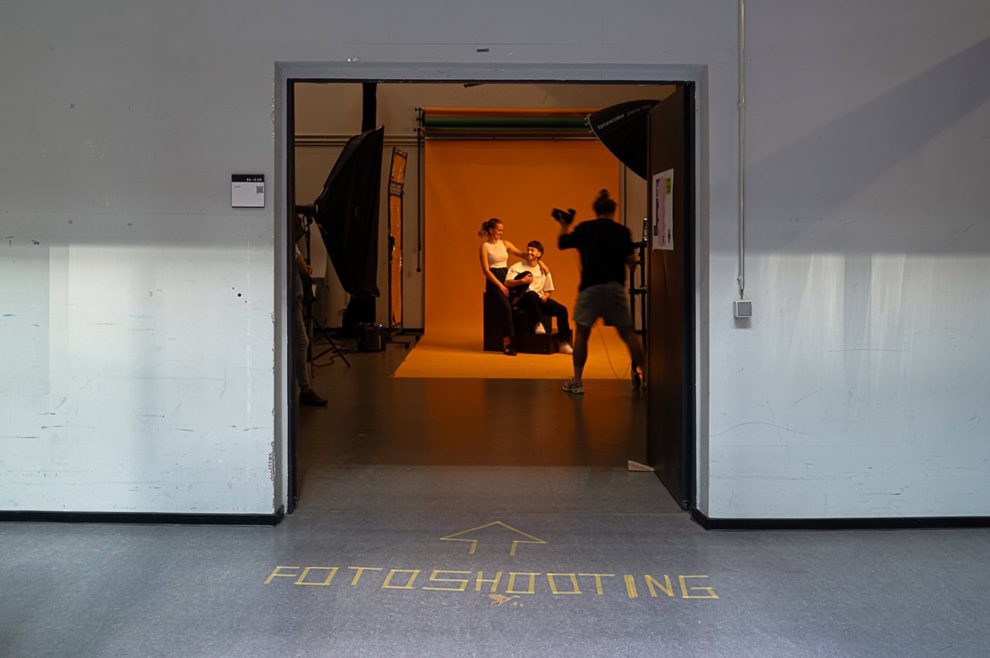 Fotografie-Studierende während einer Fotosession in einem Studio mit Beleuchtungsausrüstung und Hintergrund. Ein Schild auf dem Boden zeigt ‚FOTOSHOOTING‘ und weist auf den Eingang zum Studio hin. Von hinten sind die Umrisse eines jungen Mannes zu sehen, der Fotografie studiert. 