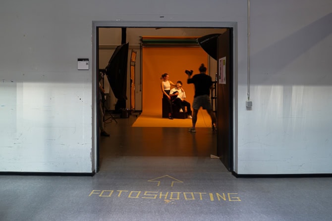 Fotografie-Studierende während einer Fotosession in einem Studio mit Beleuchtungsausrüstung und Hintergrund. Ein Schild auf dem Boden zeigt ‚FOTOSHOOTING‘ und weist auf den Eingang zum Studio hin. Von hinten sind die Umrisse eines jungen Mannes zu sehen, der Fotografie studiert.