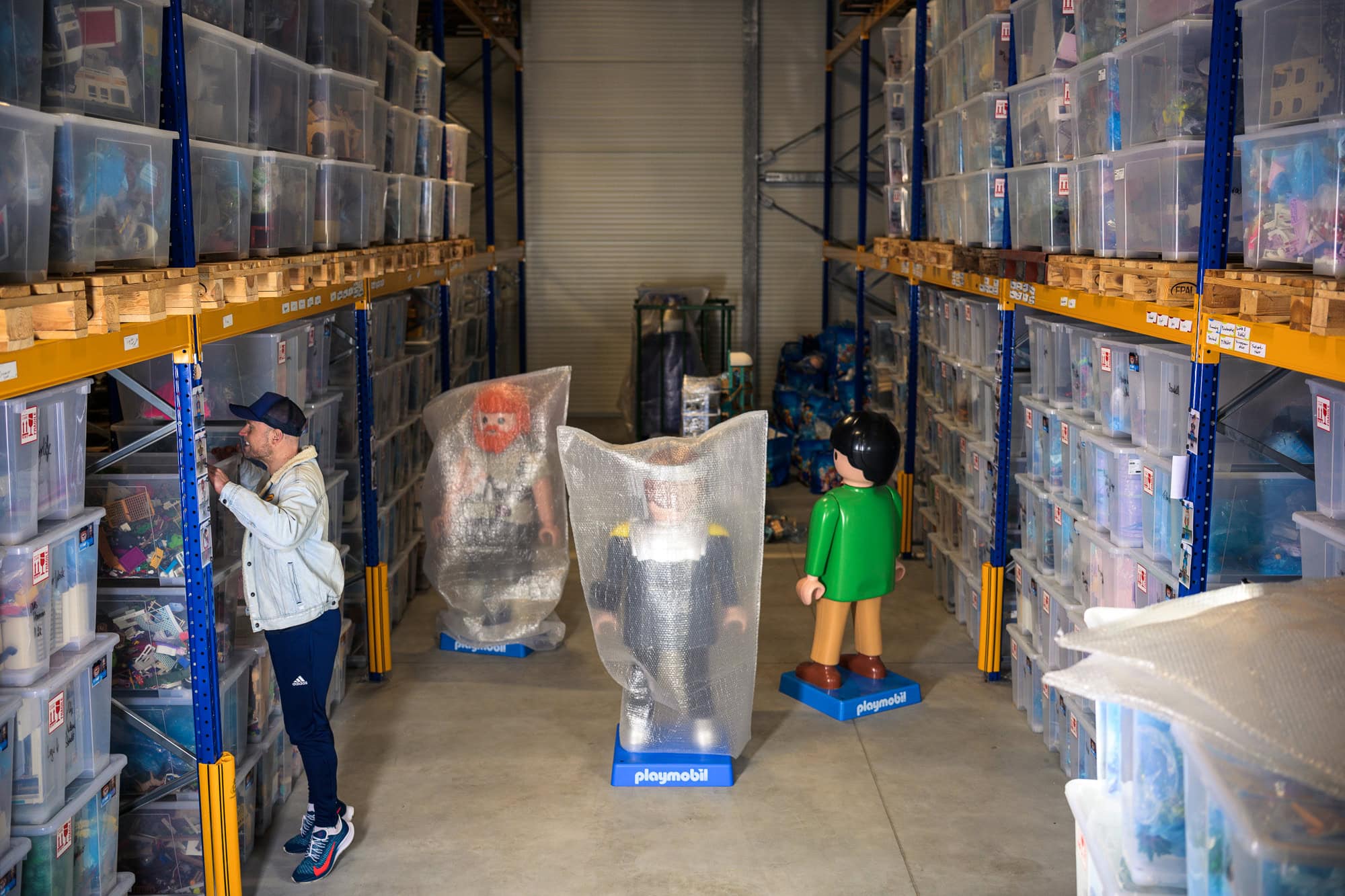 Playmobil Diorama Künstler Oliver Schaffer sucht in seinen Kisten im Hochregal Lager. Im Hintergrund stehen lebensgroße Playmobil Figuren, die teilweise in Luftpolsterfolie eingewickelt sind.