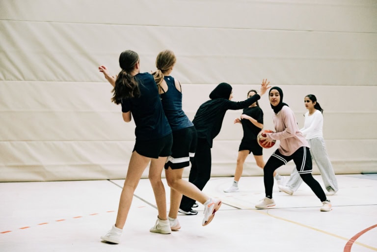Mehrere junge Frauen spielen in einer Turnhalle Basketball. Eine Spielerin im Vordergrund hält den Ball und bewegt sich nach vorne, während andere Spielerinnen sie verteidigen und die Arme heben. Die Gruppe trägt sportliche Kleidung in Schwarz, Weiß und hellen Farben. Der Moment zeigt Bewegung, Konzentration und Teamspiel in einer hellen Indoor-Sporthalle.