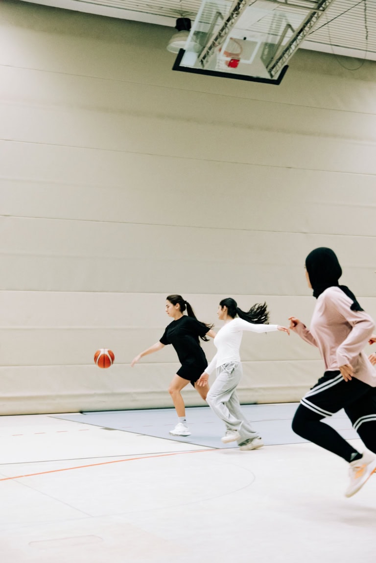 Drei junge Frauen bewegen sich dynamisch über den Hallenboden einer Turnhalle. Eine Spielerin links dribbelt einen Basketball nach vorne, während zwei andere Spielerinnen ihr dicht folgen und versuchen, sie einzuholen. Die Szene zeigt schnelle Bewegung, sportliche Anstrengung und Wettkampf in einer hellen Indoor-Sporthalle.
