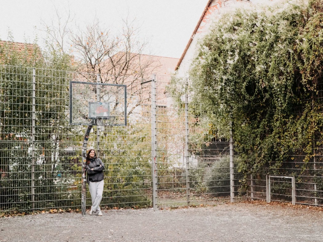 Die FLINTA Person Janne, die Gründerin der Ball like a Girl Basketball Gruppe, lehnt an dem Bein des Basketballkorb auf dem Freiplatz.