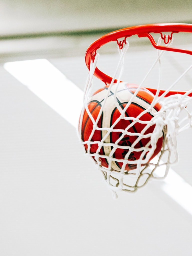 Ein Basketball fällt durch den roten Ring eines Korbes und spannt dabei das weiße Netz. Das Bild zeigt den Moment des erfolgreichen Wurfs aus der Nähe, mit Fokus auf Ball, Netz und Korb vor hellem Hintergrund.