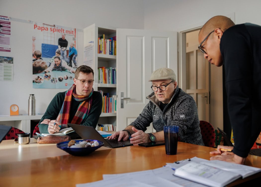 Drei Männer unterschiedlichen Alters arbeiten gemeinsam an einem Tisch in einem Büro oder Besprechungsraum. In der Mitte sitzt ein älterer Mann mit Brille, grauer Schiebermütze und Strickpullover, der mit ernster Miene auf einen Laptop zeigt. Links neben ihm sitzt ein jüngerer Mann mit Brille und bunt karierter Wollschal, der aufmerksam zuhört und Notizen macht. Rechts beugt sich ein dritter Mann mit rasiertem Kopf und schwarzem Pullover über den Tisch und blickt konzentriert auf den Bildschirm. Der Tisch ist mit einem blauen Glas, einem Notizbuch, einer Federmappe, einer Thermoskanne und einem Teller mit Gebäck belegt. Im Hintergrund stehen Regale voller Bücher sowie ein Plakat mit dem Titel „Papa spielt mit!“, das Väter mit Kindern beim Spielen zeigt.