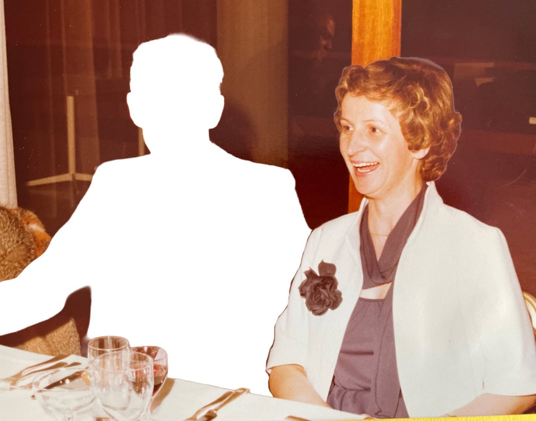 A couple sit side by side at a long table. The woman, smartly dressed, laughs past the camera. Only the man's silhouette is visible, filled with a white shape.