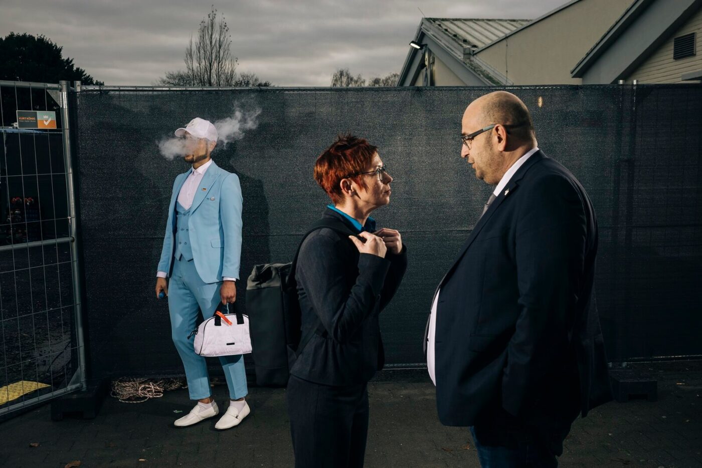 Drei Personen vor einem Bauzaun, eine Figur im hellblauen Anzug, aus der Serie Democracy Dies in Daylight