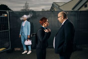Drei Personen vor einem Bauzaun, eine Figur im hellblauen Anzug, aus der Serie Democracy Dies in Daylight