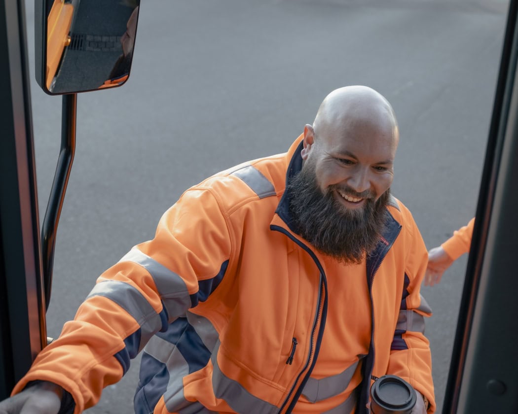 Ein lächelnder Mann mit Vollbart und Warnjacke steigt in ein Fahrzeug.
