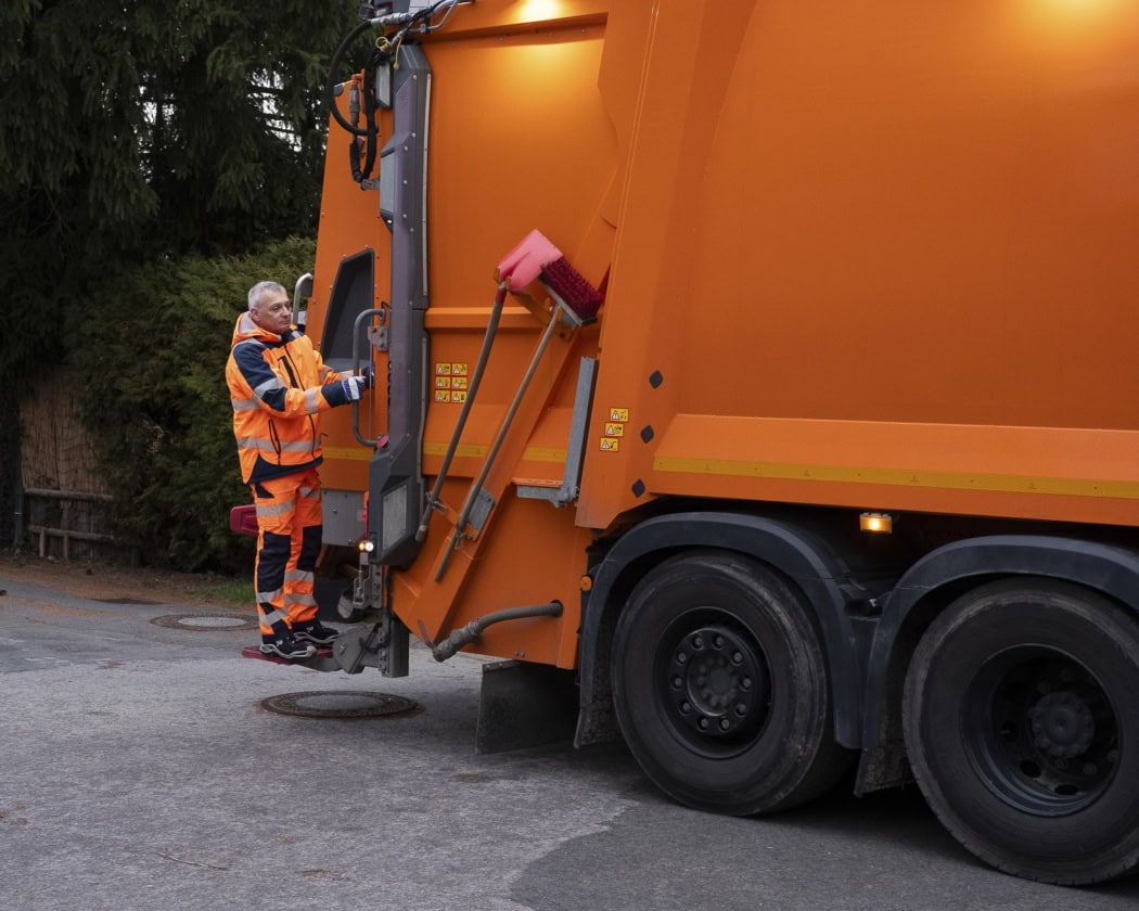 Ein Müllwerker in orangefarbener Arbeitskleidung steigt hinten auf einen Müllwagen.