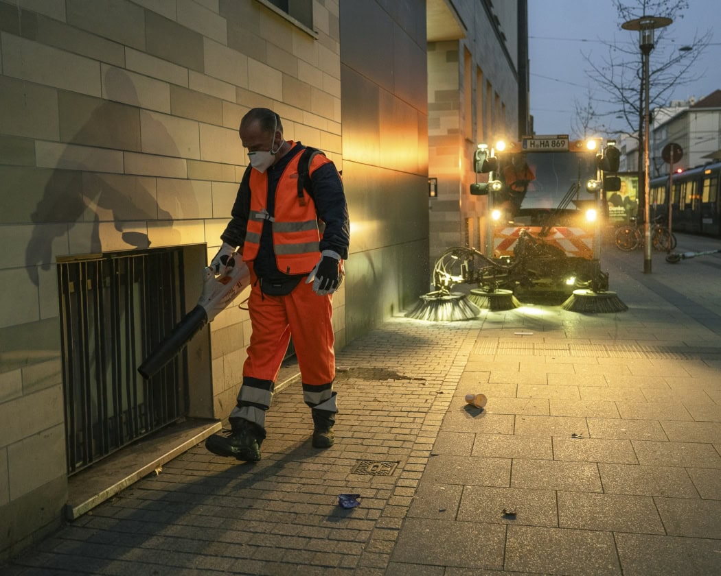 Ein Straßenreiniger mit Laubbläser auf einem Gehweg, dahinter eine Kehrmaschine im Morgenlicht.