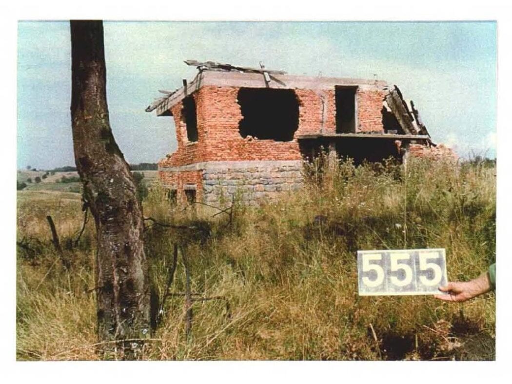 Zerstörtes Backsteinhaus auf einer Wiese, davor eine Hand mit einem Zettel mit der Nummer 555