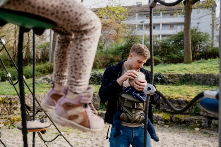 alleinerziehender Vater mit Kind. Ein Mann rückt einem Baby die Mütze zurecht, während das Baby in einer nach vorne gerichteten Babytrage an seiner Brust befestigt ist. Sie befinden sich auf einem Spielplatz, auf dem ein anderes Kind im Vordergrund zu sehen ist, das auf einem Seil klettert und rosa Turnschuhe und Leggings mit Leopardenmuster trägt. Im Hintergrund sind Grünflächen und Wohngebäude zu sehen, die an einen Stadtpark erinnern.