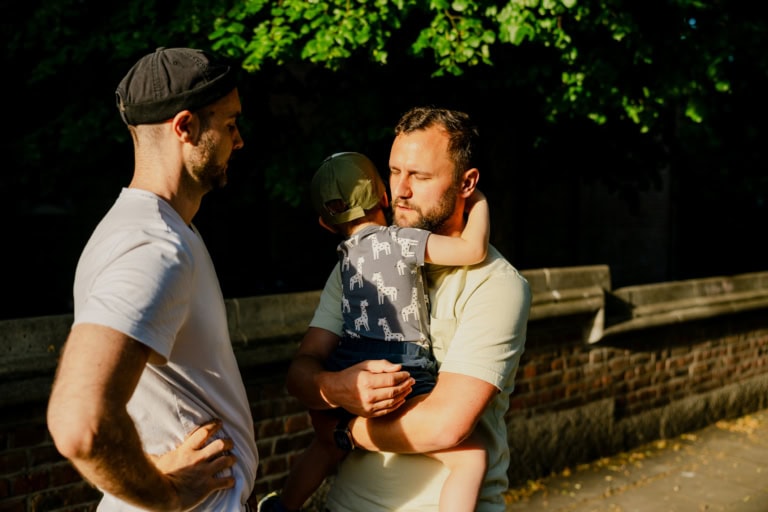 moderne Familienform, Zwei Männer stehen im Freien in weichem, goldenem Sonnenlicht; ein Mann, der ein helles Hemd trägt, hält ein Kind, das sich an ihn klammert. Das Kind, das ein giraffengemustertes Hemd und eine Mütze trägt, hat seine Arme um den Hals des Mannes geschlungen. Der zweite Mann, mit weißem Hemd und Hut, sieht zu, wie sie in der Nähe einer Backsteinmauer stehen, die im Hintergrund begrünt ist.