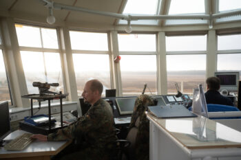 Ein Soldat in Tarnuniform arbeitet im Kontrollraum der Nordhorn Range, einer militärischen Schießanlage, vor einem Computer. Ein weiterer Soldat ist im Hintergrund an einem anderen Arbeitsplatz zu sehen, während der Kontrollraum durch große Fenster den Blick auf die Anlage und die umliegende Landschaft bietet.