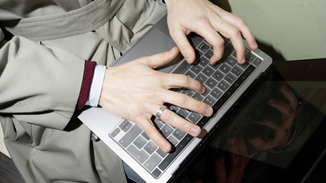 Das Bild zeigt eine Nahaufnahme von zwei Händen, die auf der Tastatur eines Laptops tippen. Die Person trägt einen beigefarbenen Mantel und darunter ein hellblaues Hemd mit einem weinroten Pullover. Ein silberner Ring schmückt einen Finger der rechten Hand. Der Laptop liegt auf dem Schoß der Person, und die Reflexion der Hände ist teilweise auf der glänzenden Oberfläche des Geräts sichtbar. Die Szene wirkt dynamisch und konzentriert.