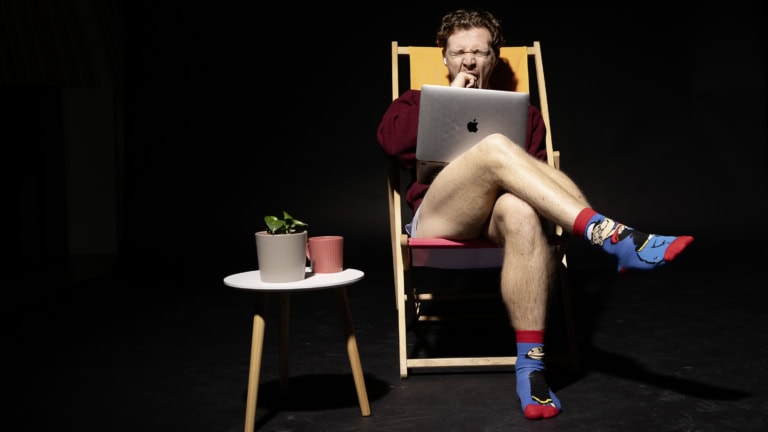 Das Bild zeigt einen jungen Mann, der in einem Liegestuhl sitzt, während er auf einem Laptop arbeitet. Er trägt ein weinrotes Oberteil und auffällige bunte Socken mit Comic-Motiven, ist jedoch sonst leger gekleidet. Auf einem kleinen weißen Beistelltisch neben ihm stehen eine grüne Topfpflanze und eine rosa Tasse. Der Hintergrund ist dunkel, wodurch die Szene und die Gegenstände im Vordergrund hervorgehoben werden. Er wirkt entspannt, ist jedoch am gähnen, was möglicherweise auf Müdigkeit hinweist.