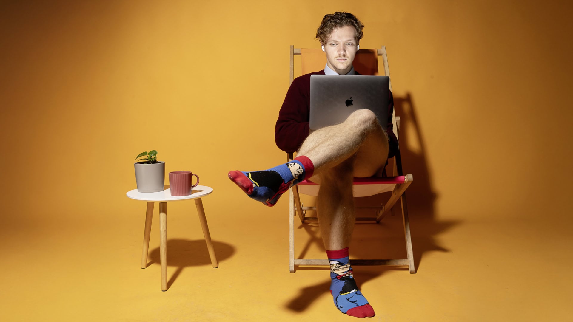 Ein junger Mann mit hellbraunen Locken sitzt auf einem Klappstuhl. Der Hintergrund ist leuchtend orange. Er trägt einen weinroten Pullover, darunter ein hellblaues Hemd. Er trägt keine richtige Hose, aber bunte Socken mit Comic-Motiv. Seine Beine hat er überschlagen. Neben ihm steht ein weißer dreibeiniger Couchtisch, auf dem ein grauer Blumentopf mit einer Pflanze und eine rosa Tasse stehen. Er schaut konzentriert auf seinen Laptop auf seinem Schoß. In seinen Ohren hat er Bluetooth-Kopfhörer.