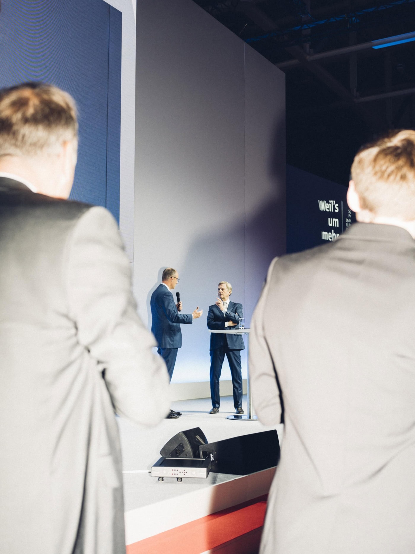 CDU-Vorsitzender Friedrich Merz mit Mikrofon in der Hand, im Gespräch mit dem ehemaligen ZDF-Moderator Klaus Kleber auf einer Bühne. Im Vordergrund sind zwei Männer angeblitzt von hinten zu sehen.