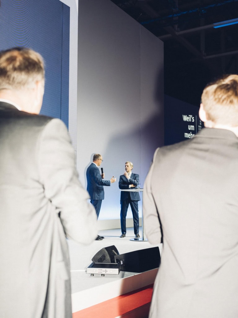 CDU-Vorsitzender Friedrich Merz mit Mikrofon in der Hand, im Gespräch mit dem ehemaligen ZDF-Moderator Klaus Kleber auf einer Bühne. Im Vordergrund sind zwei Männer angeblitzt von hinten zu sehen.
