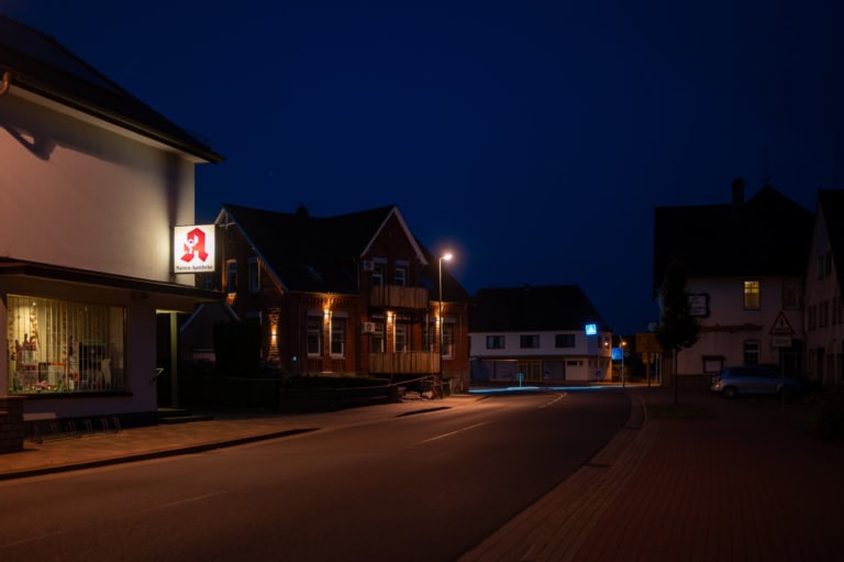 Auf dem Foto ist eine Dorfstraße zu sehen. Es ist Nacht und die Straßenlaternen leuchten. Auf der linken Seite ist eine beleuchtete Apotheke zu sehen.
