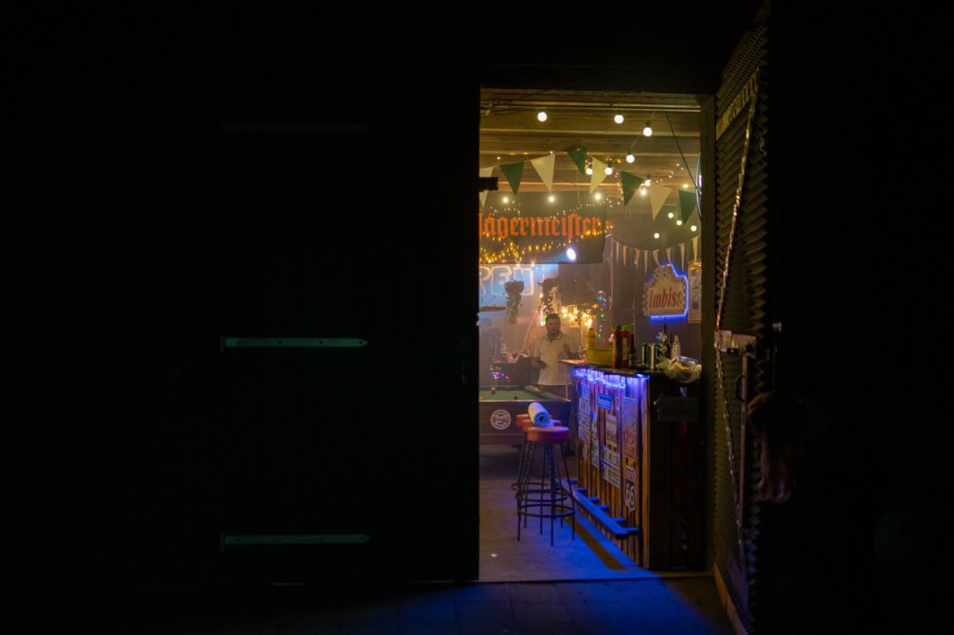 Auf dem Foto ist eine geöffnete Scheunentür zu sehen. Draußen ist es dunkel. Der Innenraum der Scheune ist mit Lichterketten und Partylicht erleuchtet. Es ist ein Bartresen, ein Billardtisch und Jägermeister-Werbung zu sehen.