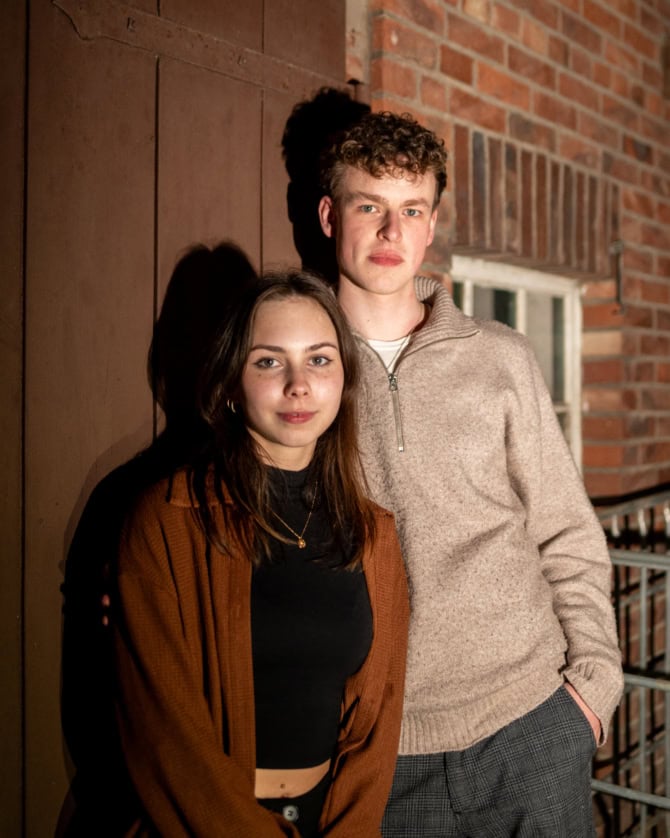 Auf dem Foto sind ein junger Mann und eine junge Frau zu sehen. Sie schauen in die Kamera. Sie stehen vor einer Scheune. Es ist Nacht.