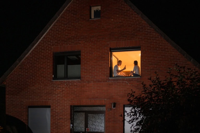 Auf dem Foto ist ein rotes Backsteinhaus zu sehen. Es ist Nacht. An einem erleuchteten Fenster sitzen zwei Personen auf der Fensterbank. Die linke Person prostet der rechten Person mit einer Flasche zu.