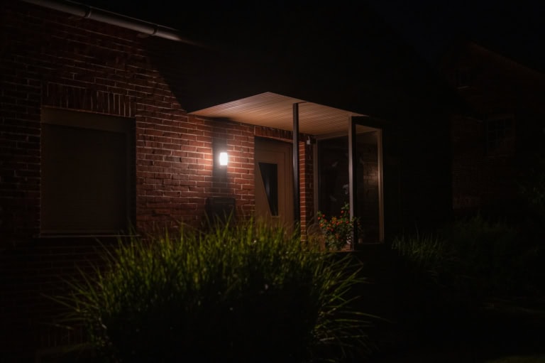 Auf dem Foto ist ein Hauseingang eines Backsteinhauses zu sehen. Es ist nachts und dunkel. Eine kleine Lampe leuchtet neben der Eingangstür.