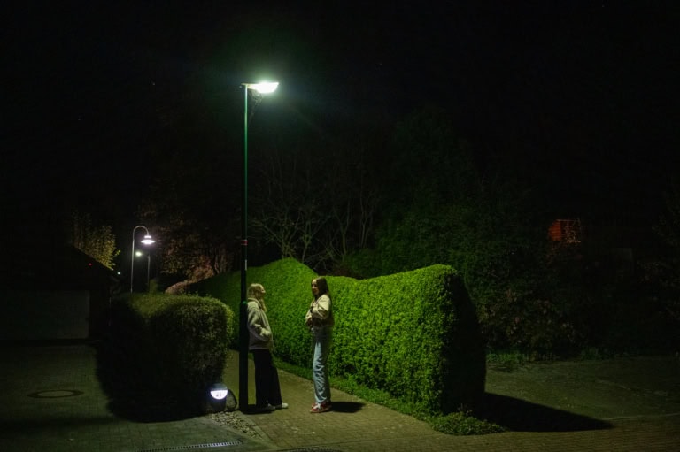 Auf dem Foto sind zwei Personen zu sehen die unter einer Straßenlaterne, zwischen hohen Hecken stehen und sich unterhalten. Es ist Nacht.
