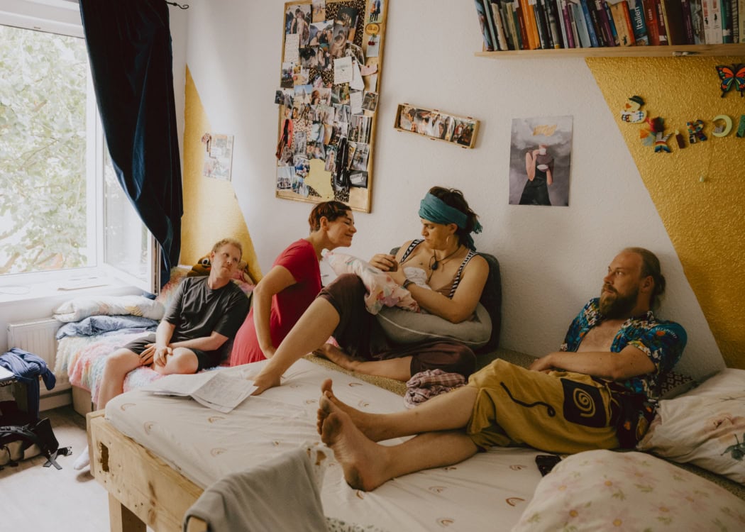 Mutter Julia stillt ihr Kind. Eine Hebamme und ihr Kumpel Ingmar (links), mit dem sie in einem Co-Parenting Konzept lebt, schauen dem Kind zu. Rechts schaut ihr Beziehungspartner Patrick zu Verle