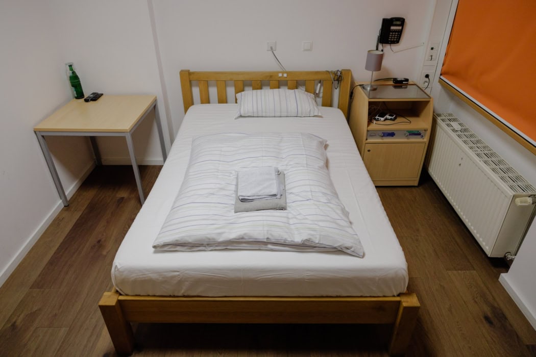 Schlicht eingerichtetes Einzelzimmer mit einem Holzbett, das mit weißer Bettwäsche bezogen ist, und einem gefalteten Handtuch auf der Bettdecke. Neben dem Bett steht ein Nachttisch mit einer Lampe, einem Telefon und einigen Geräten, während ein kleiner Schreibtisch mit einer Wasserflasche und Fernbedienungen im Hintergrund sichtbar ist. Das Zimmer befindet sich in der Advanced Sleep Research GmbH und wird für die Schlafüberwachung genutzt.